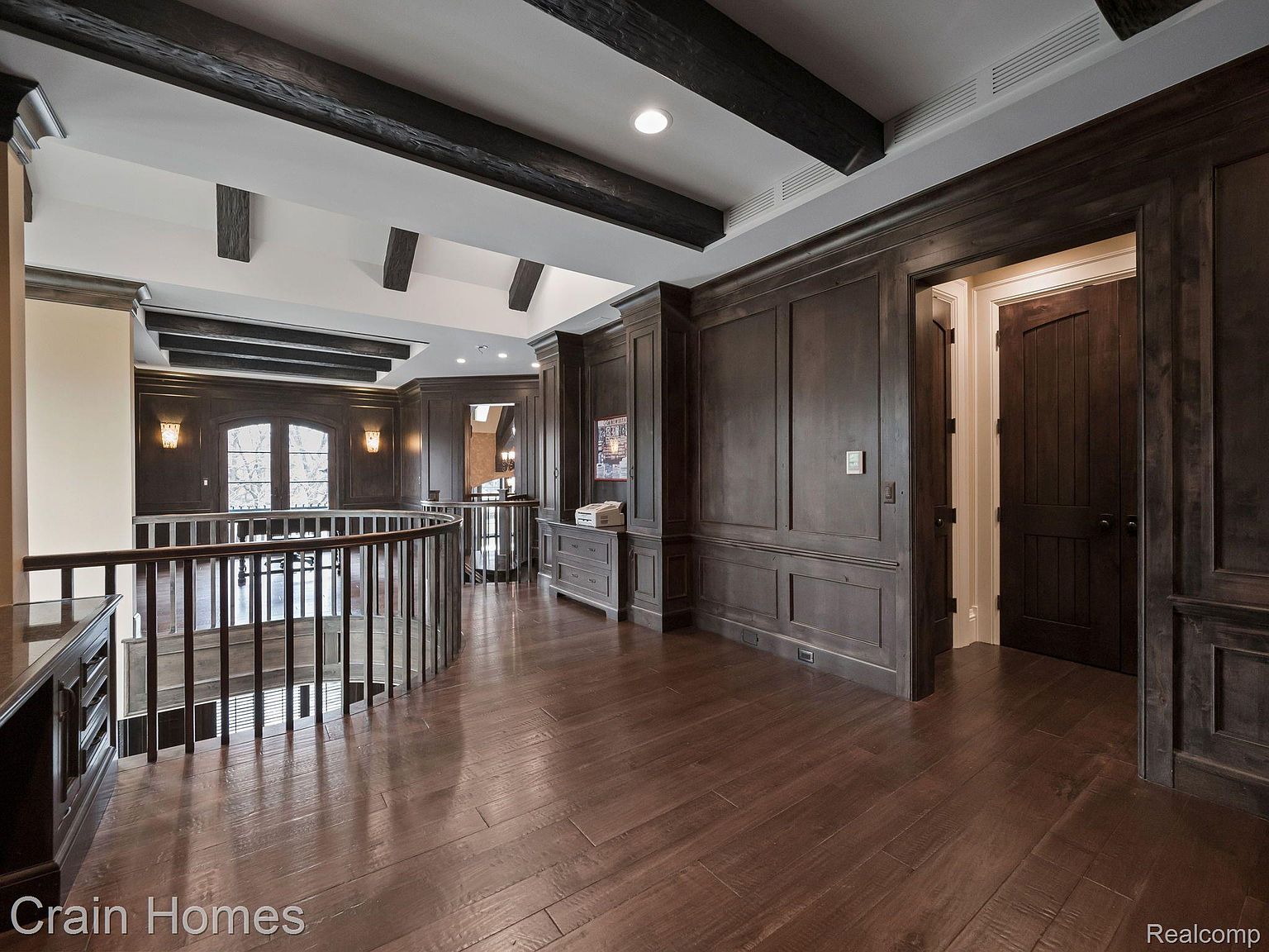 This interior shot showcases a luxurious hallway with dark wood flooring and paneled walls, creating a sophisticated and elegant atmosphere. A curved staircase with dark wood railings adds a focal point, while the dark wood beams on the ceiling contribute to the room's architectural interest. The space is well-lit, highlighting the rich textures and finishes.