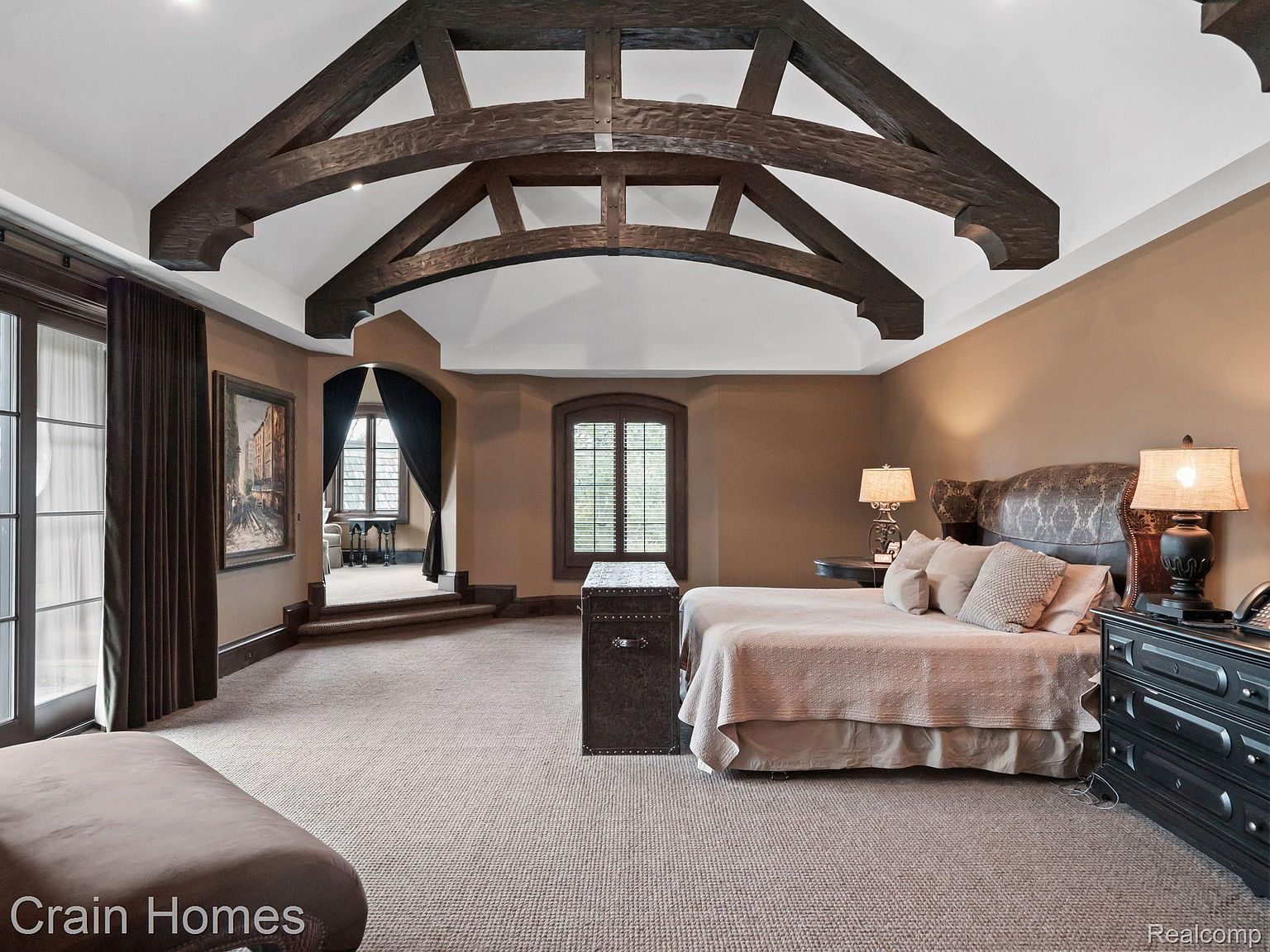 This is a spacious primary bedroom featuring a unique architectural ceiling with exposed wooden beams. The room is decorated in warm, earthy tones, with a large bed, dark wood furniture, and a separate seating area visible through an arched doorway. The overall impression is luxurious and inviting.