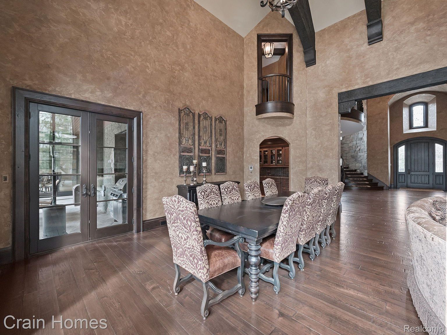 This is an interior shot of a formal dining room. The room features a large wooden dining table with upholstered chairs, set on dark hardwood floors. The walls are textured in a warm, earthy tone, complemented by dark wood trim and accents. A balcony overlooks the dining area, adding to the grandeur of the space.
