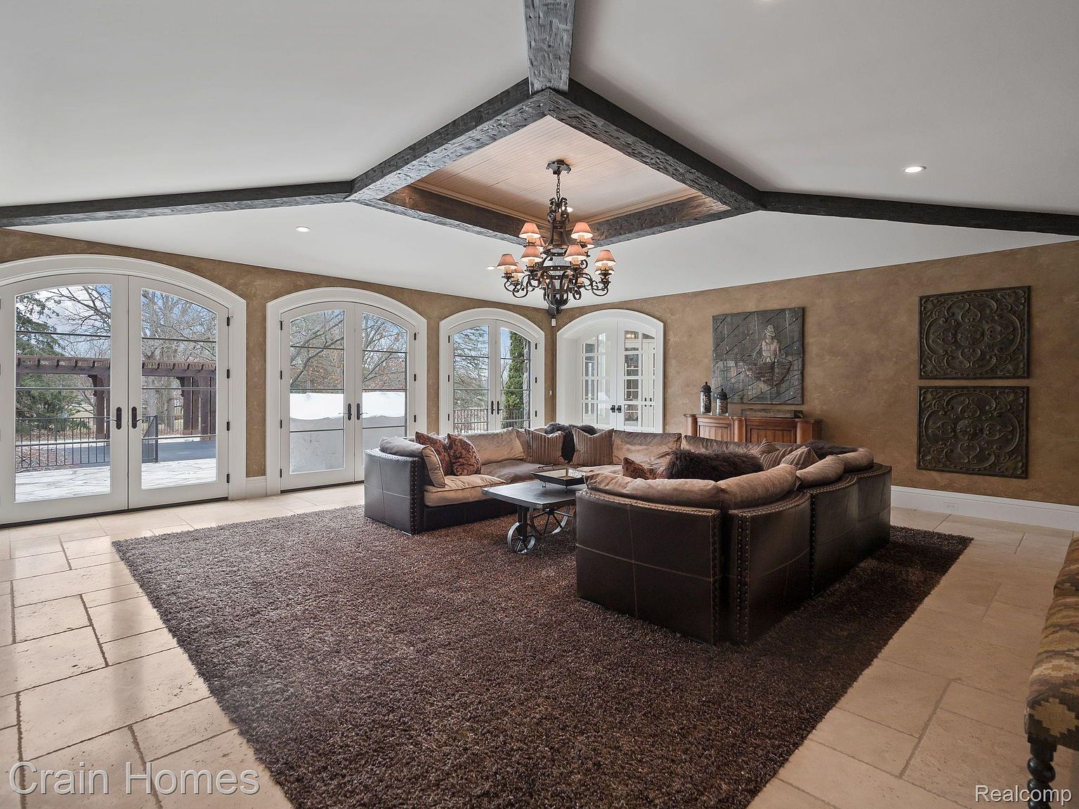 This is an interior shot of a luxurious living room featuring a large, sectional sofa with brown leather and plush cushions, centered around a dark-toned coffee table. The room is illuminated by an ornate chandelier and natural light streaming through arched French doors, complemented by a dark brown accent wall and decorative wall art. The flooring is a combination of tile and a large area rug, creating a warm and inviting atmosphere.
