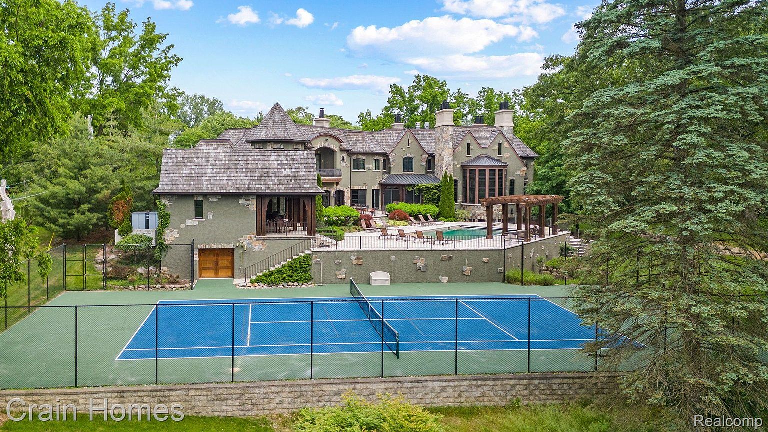 This image showcases the rear exterior of a luxurious home, complete with a tennis court, swimming pool, and meticulously landscaped grounds. The architectural style features a blend of stone and wood, creating a sophisticated and inviting atmosphere. The property exudes a sense of privacy and upscale living, perfect for entertaining and relaxation.