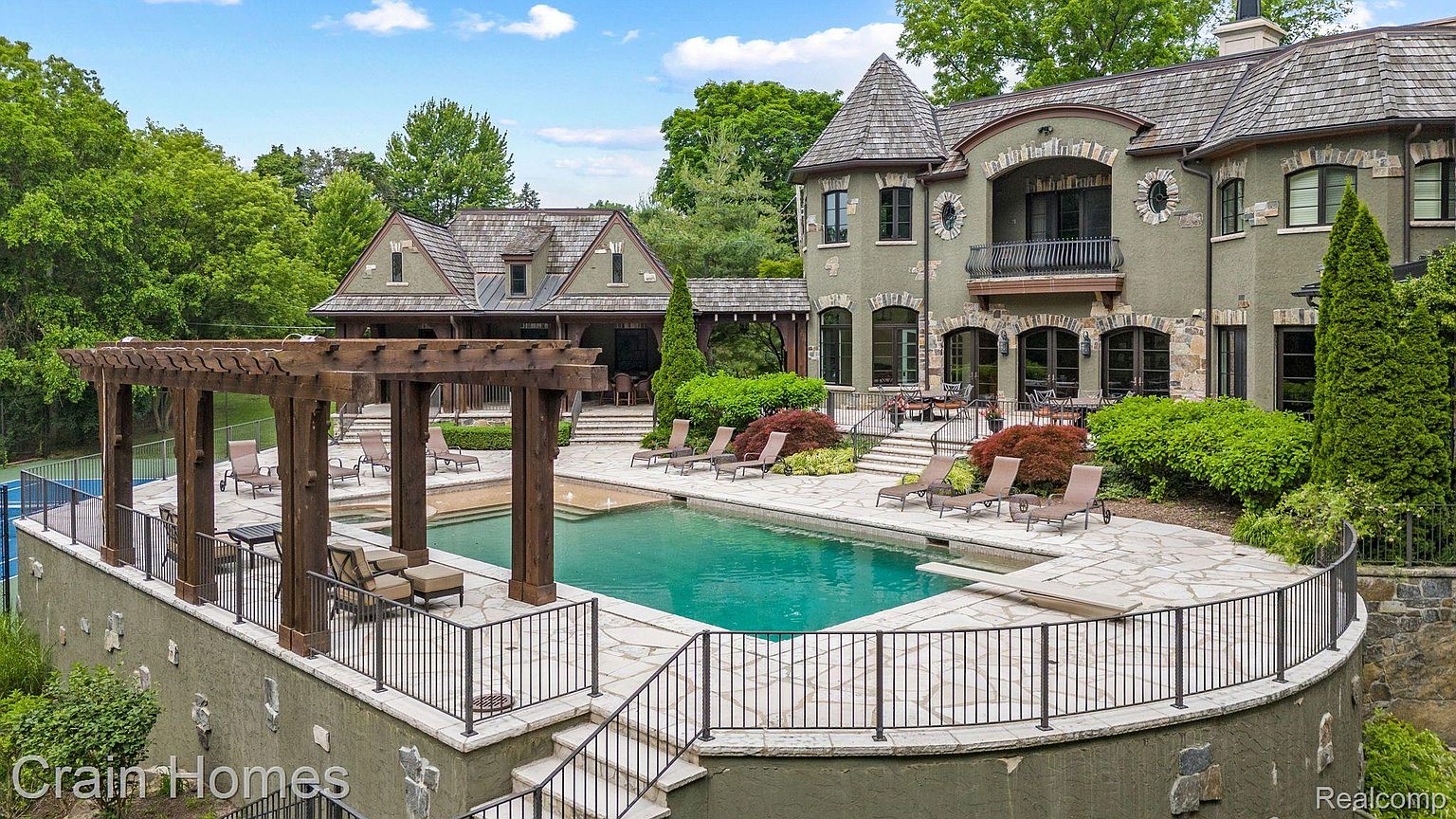 This is a rear exterior view showcasing a luxurious pool and spa area of a large estate. The pool is surrounded by a stone patio with lounge chairs, and a wooden pergola provides shade. The main house features stone accents and a turret, adding to the property's elegant and upscale appeal.