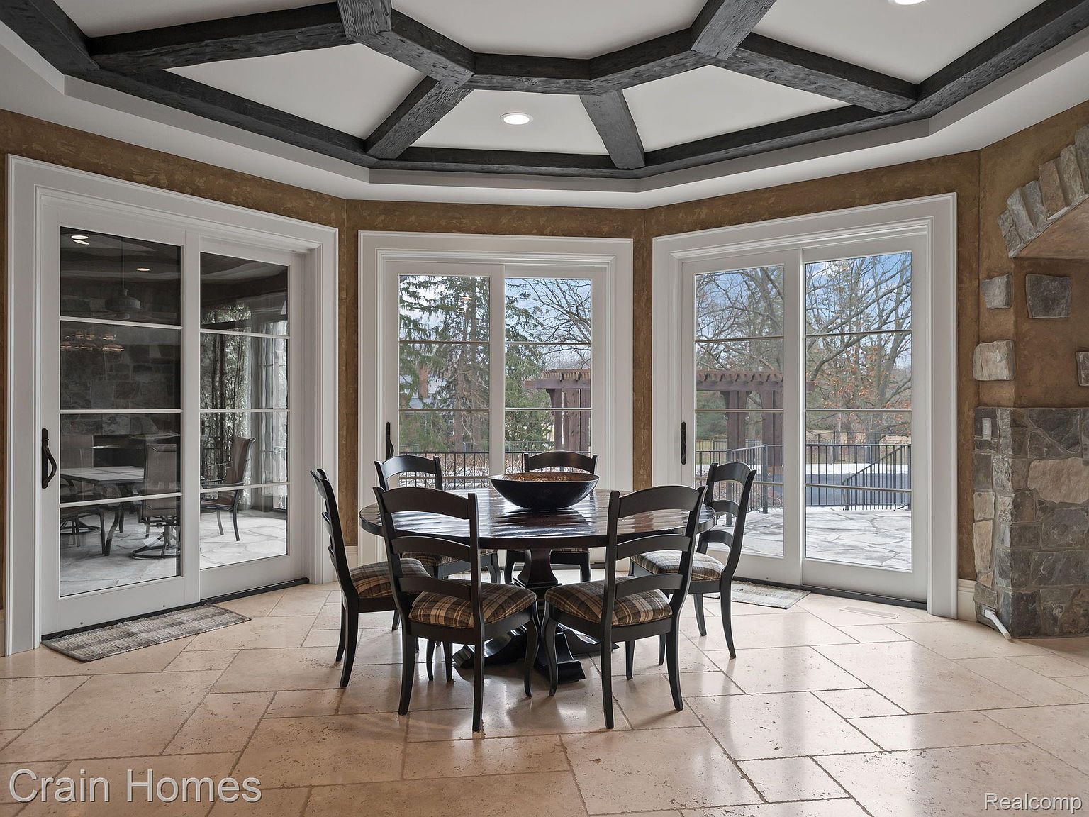 This interior shot showcases a dining room with a round wooden table surrounded by six chairs with plaid cushions. The room features a unique ceiling design with dark wooden beams and recessed lighting, complemented by large windows and a set of glass doors that offer views of the outdoor space. The flooring is tiled, adding to the room's elegant and inviting atmosphere.