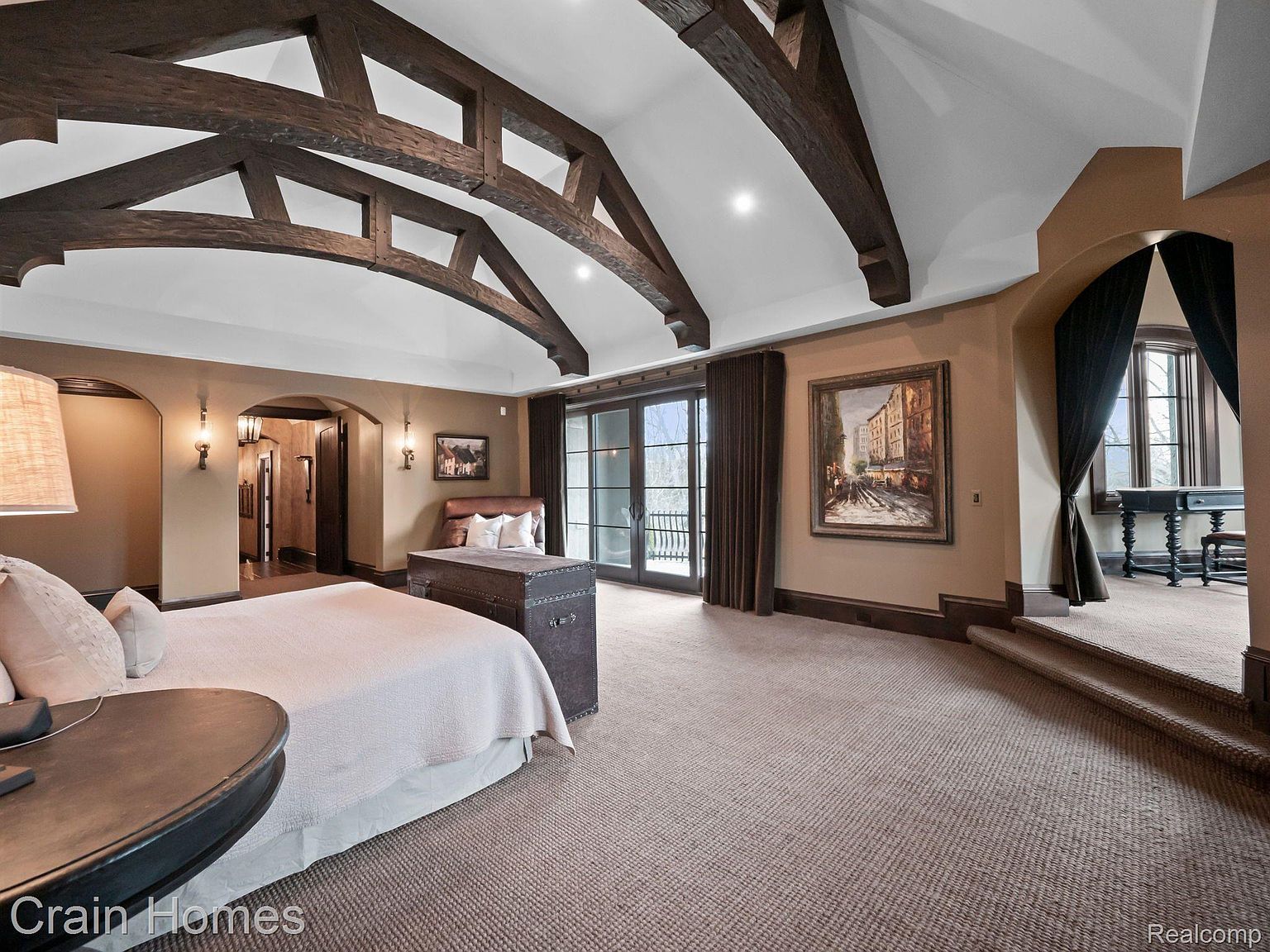 This is a luxurious primary bedroom featuring exposed wooden beams on the ceiling, a large bed with neutral bedding, and a sitting area with a trunk. The room has a warm color palette, with carpeted floors and a painting on the wall, and it leads to a raised area with a window and desk, creating a spacious and inviting atmosphere. The room also has access to an outdoor balcony.