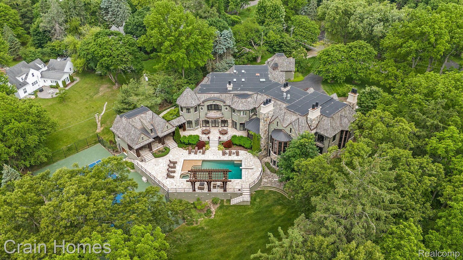 This is an aerial view of a large, luxurious estate surrounded by lush greenery. The property features a sprawling mansion with a dark roof, a swimming pool with a pergola, a tennis court, and meticulously landscaped grounds. The overall impression is one of opulence and privacy.