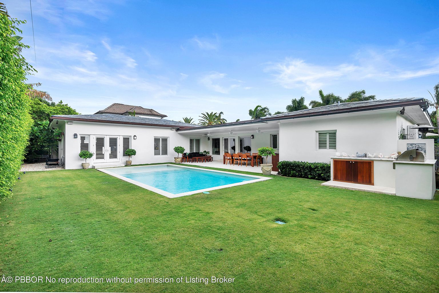 This is a rear view of a modern home featuring a well-manicured lawn, a rectangular swimming pool, and an outdoor kitchen area. The house is painted white with dark trim, and the backyard includes a patio with outdoor dining furniture. The overall impression is one of luxury and outdoor living.