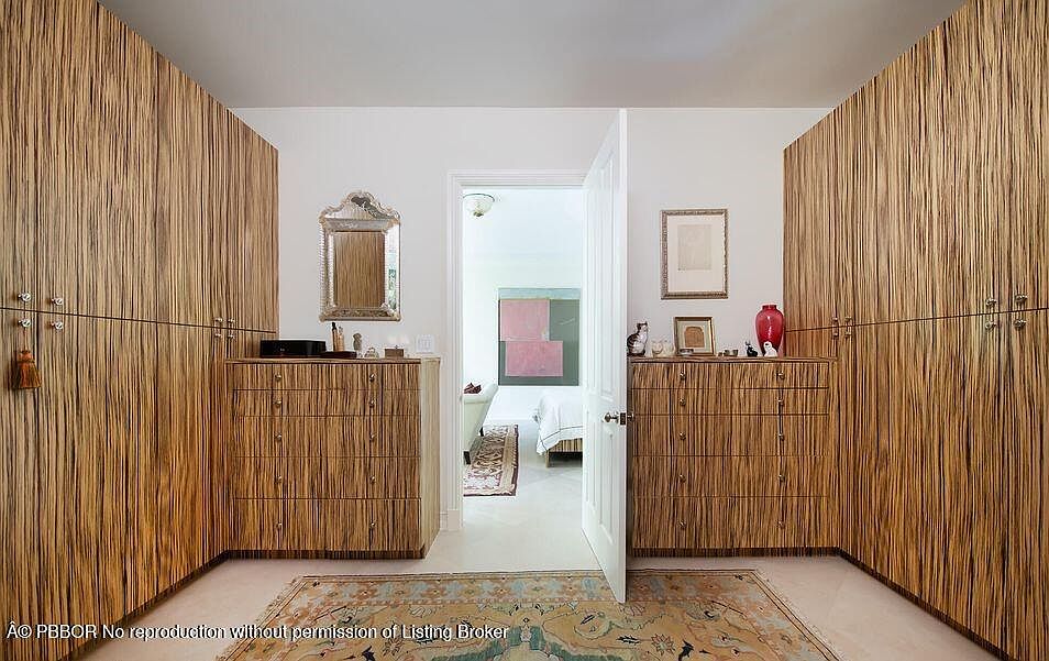 This is a well-organized walk-in closet featuring custom wood cabinetry and drawers. The closet is divided into two sections with a doorway leading to a bedroom. A decorative rug adds warmth to the space, and the overall impression is one of luxury and functionality.