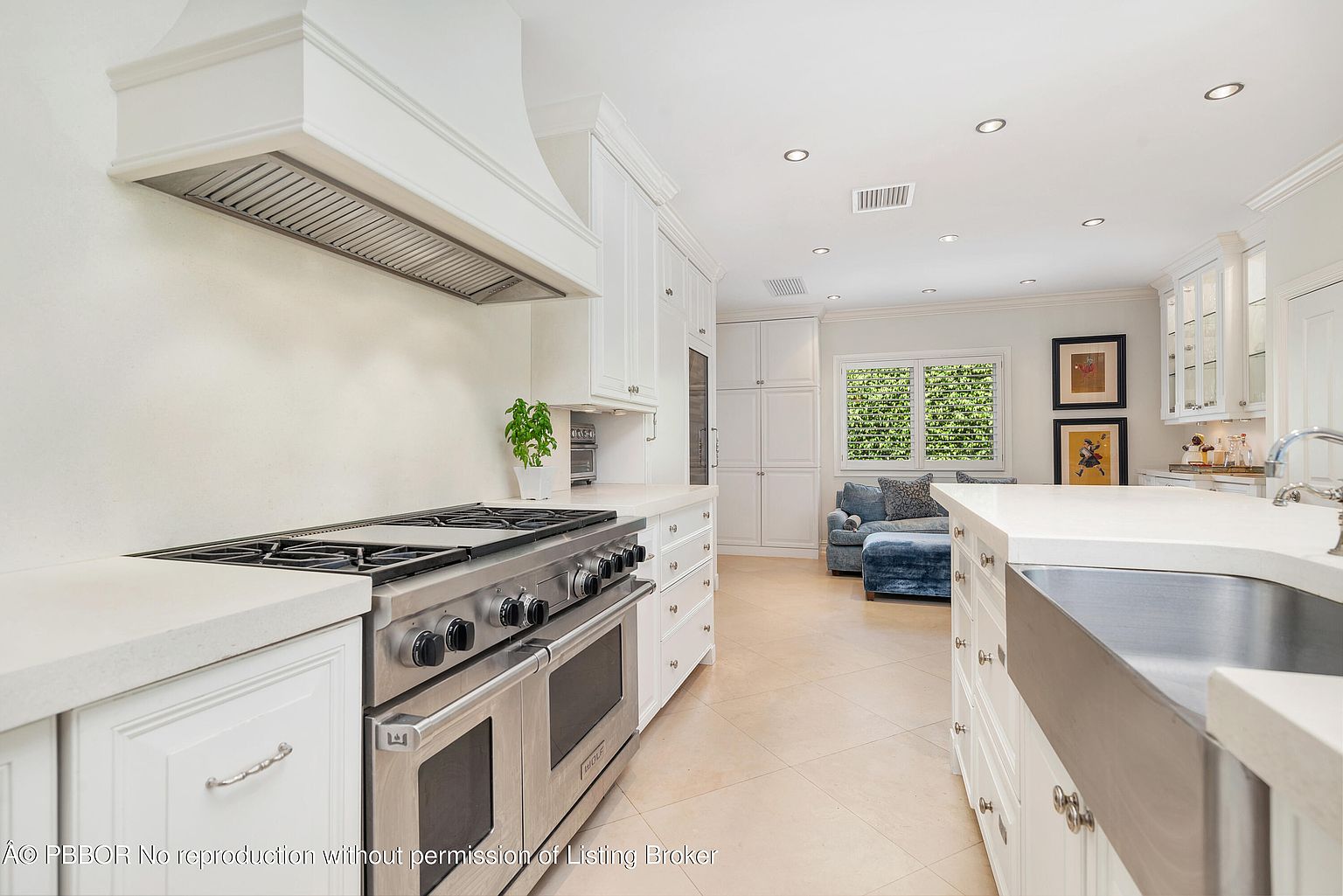 This is a bright and spacious kitchen featuring white cabinetry, stainless steel appliances, and light-colored countertops. A large range hood is positioned above the stove, and the kitchen opens into a living area with a blue sofa. The flooring is a light tile, and the overall impression is clean and modern.