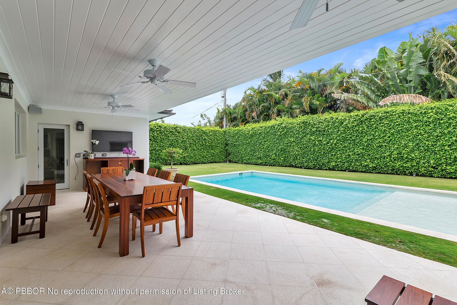This image showcases a covered patio area adjacent to a swimming pool. The patio features a dining table with wooden chairs, a TV mounted on the wall, and ceiling fans for comfort. The pool is surrounded by a well-maintained lawn and a tall, manicured hedge, providing privacy and a lush backdrop.