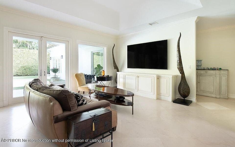 This is an interior shot of a living room featuring a tan leather sofa with brown pillows, a dark wood coffee table, and a dark wood chest on a stand. A large flat-screen TV is mounted on the wall above a built-in white cabinet. The room has a neutral color palette with light-colored flooring and walls, creating a bright and airy atmosphere.