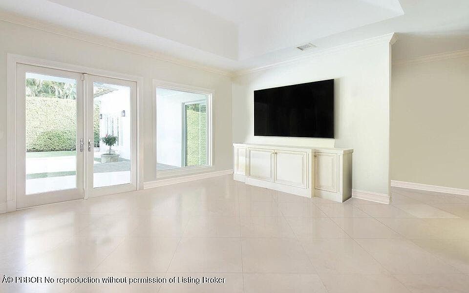 The image showcases a bright and spacious living room with a minimalist design. The room features large sliding glass doors leading to an outdoor area, a window, and a wall-mounted television above a white cabinet. The flooring is light-colored tile, contributing to the room's airy and modern aesthetic.