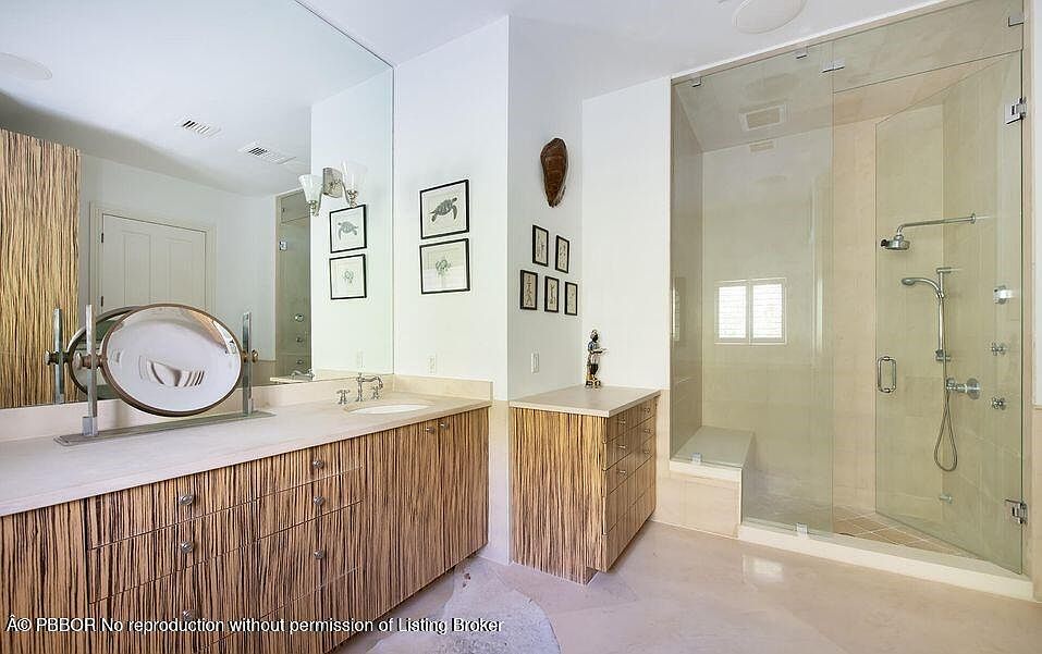 This is a primary bathroom featuring a modern design with a large mirror, custom wood-grain cabinetry, and a glass-enclosed shower. The countertops are light-colored, and the overall aesthetic is clean and luxurious. The shower has a built-in bench and multiple shower heads.