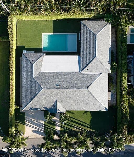 This aerial shot showcases a well-maintained property featuring a house with a gray shingle roof, a rectangular swimming pool, and lush green landscaping. The house has a complex roofline with multiple gables and a white section, suggesting a modern architectural style. The surrounding greenery provides privacy and enhances the property's appeal.