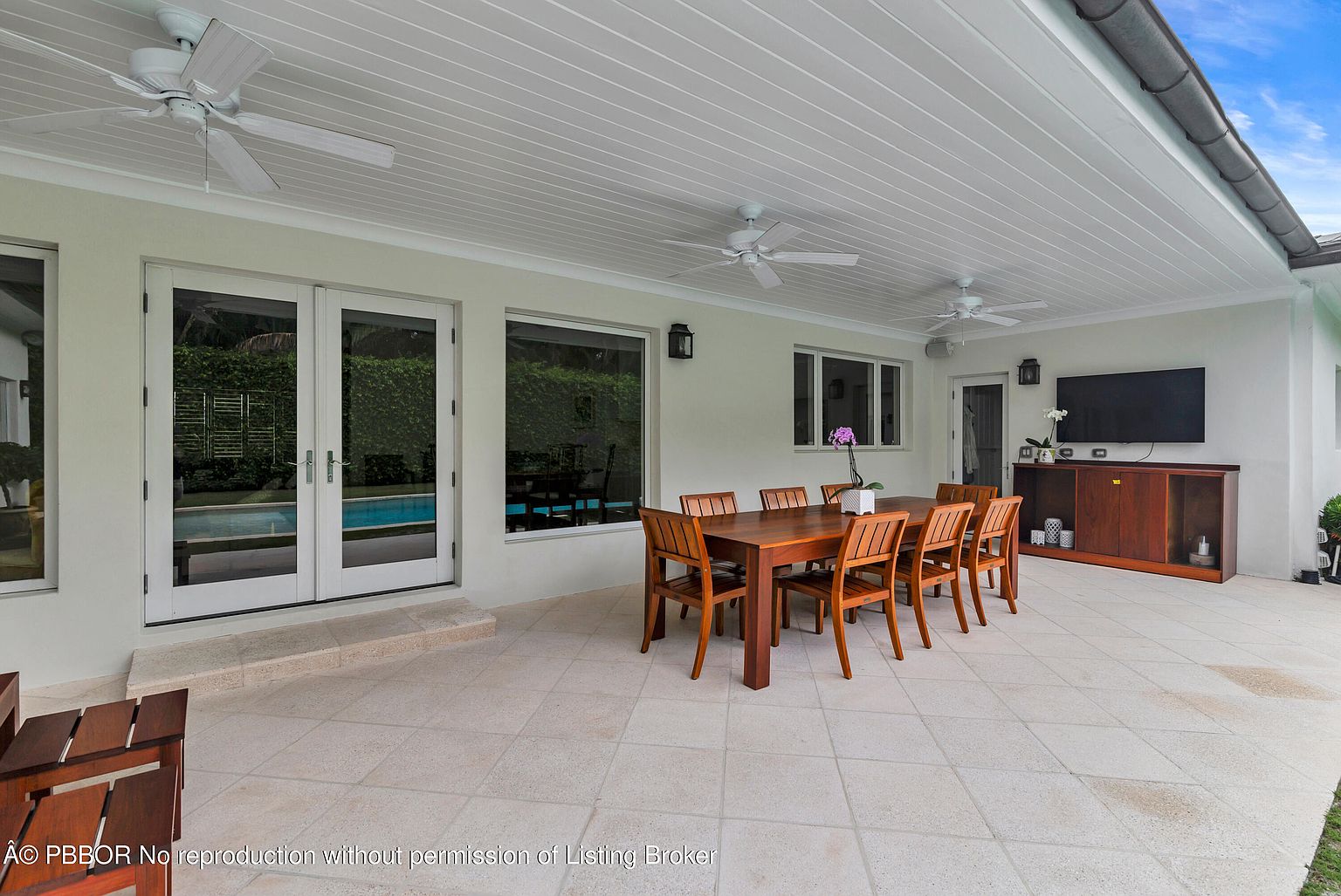 This is an exterior shot of a covered patio area, featuring a large wooden dining table with chairs, perfect for outdoor entertaining. The patio is paved with light-colored tiles, and the space is well-lit with ceiling fans and recessed lighting. The area includes a built-in cabinet with a television, enhancing the outdoor living experience.