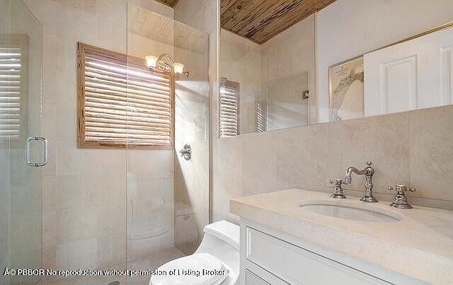 This is a primary bathroom featuring a glass-enclosed shower with a wooden-framed window, a toilet, and a vanity with a light-colored countertop and white cabinets. The walls are tiled in a neutral tone, and the overall style is clean and modern. A large mirror reflects the space, enhancing the sense of openness.