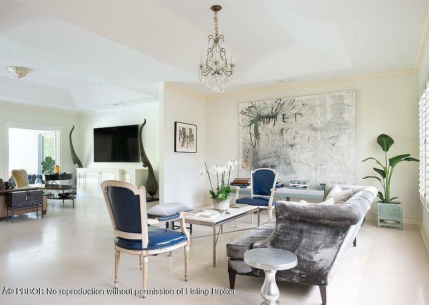 This is an interior shot of a living room featuring a chandelier, artwork, and stylish furniture. The room is bright and airy, with a neutral color palette and elegant decor. The composition includes a chaise lounge, chairs, and a coffee table, creating a sophisticated and inviting space.