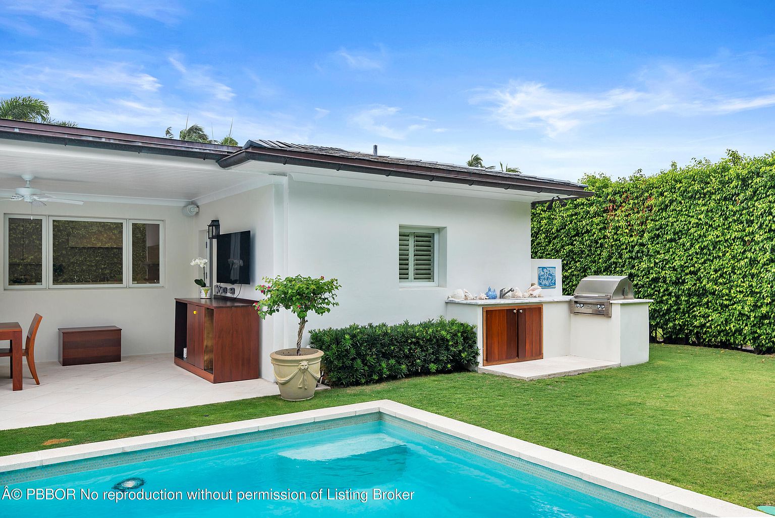 This image showcases a beautiful backyard pool area, featuring a modern outdoor kitchen with a built-in grill and cabinetry. The pool's clear blue water contrasts with the lush green lawn and manicured hedges, creating a serene and inviting atmosphere. The architectural style of the adjacent building is clean and contemporary, enhancing the overall appeal of the outdoor space.
