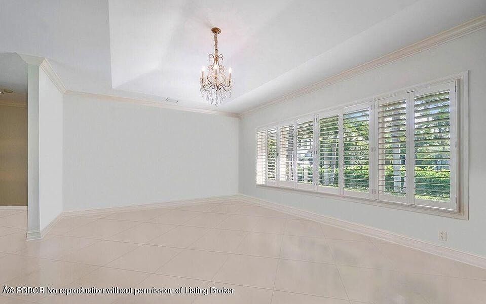 This is an interior shot of a dining room featuring a chandelier, large windows with shutters, and tile flooring. The walls are painted in a neutral color, and the room appears spacious and well-lit. The overall impression is elegant and inviting, suitable for formal dining.