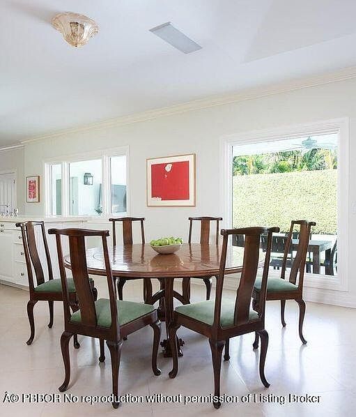 This is an interior shot of a dining room featuring a round wooden table surrounded by six chairs with green cushions. A red framed artwork hangs on the wall, and natural light streams in through a large window, creating a bright and inviting atmosphere. The room has a classic and elegant style.