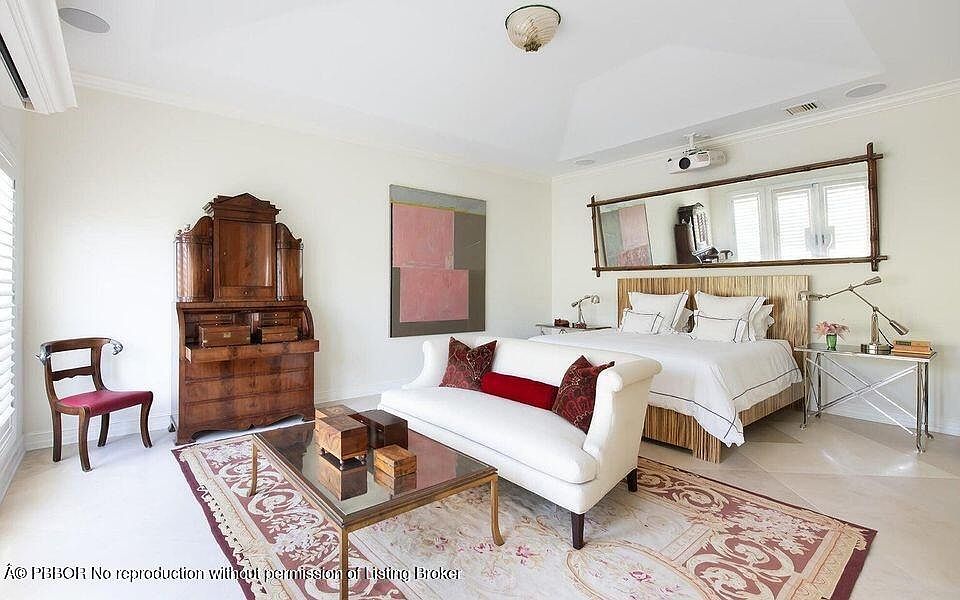 This primary bedroom features a blend of classic and contemporary styles. A white sofa with decorative pillows sits in front of a glass-topped coffee table, while a wooden secretary desk and chair are positioned to the side. The bed has a wooden headboard and is flanked by side tables, with a large mirror hanging above, and a projector is mounted on the ceiling.
