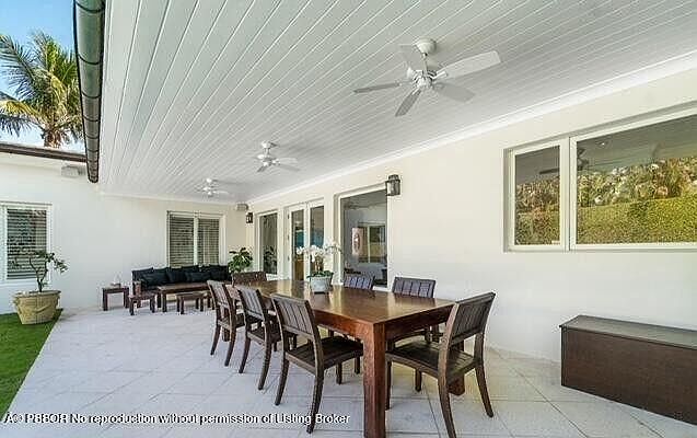 This image showcases a covered outdoor patio area, furnished with a large wooden dining table and multiple chairs, suggesting an ideal space for outdoor dining and entertaining. The patio is well-lit and features a view of the surrounding landscape through large windows, enhancing the appeal of the outdoor living space. The presence of ceiling fans indicates a focus on comfort in warmer climates.