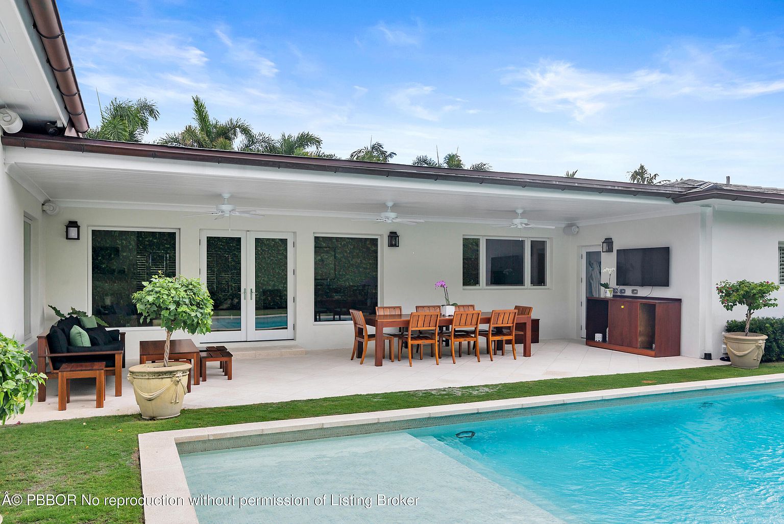 This image showcases a luxurious outdoor living space featuring a pristine pool with a shallow entry area. A covered patio area includes a dining table with chairs and an outdoor entertainment setup, creating an inviting space for relaxation and gatherings. The well-maintained lawn and landscaping add to the overall appeal of this backyard oasis.