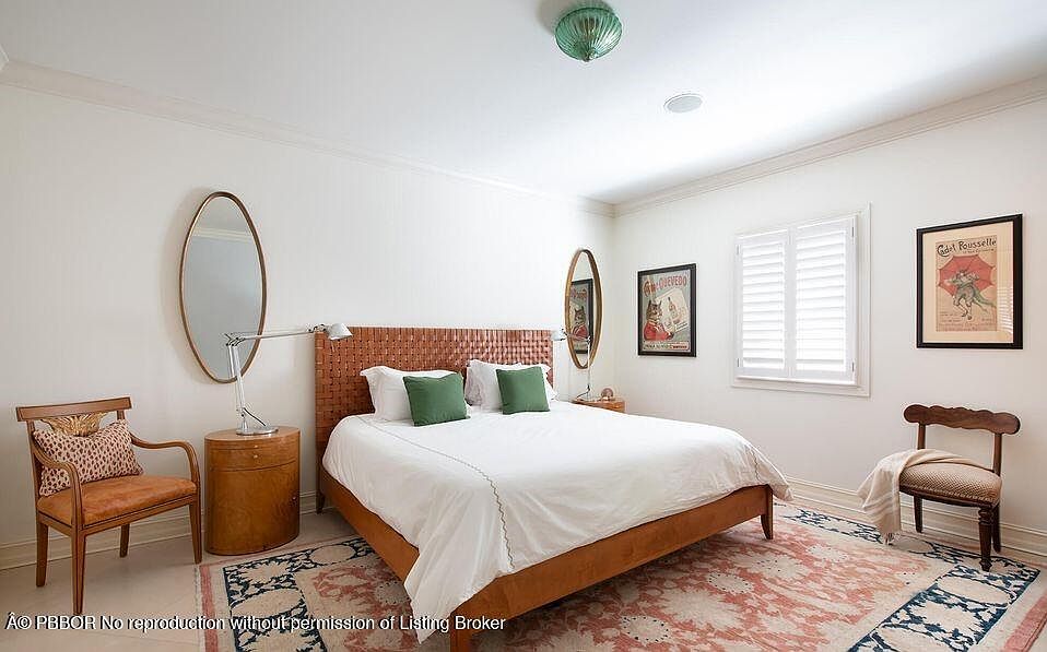 This is a well-lit bedroom featuring a bed with a woven headboard, white linens, and green pillows. The room is decorated with oval mirrors, framed artwork, and a patterned rug, creating a cozy and inviting atmosphere. The walls are painted in a neutral tone, complemented by white trim and shutters on the window.