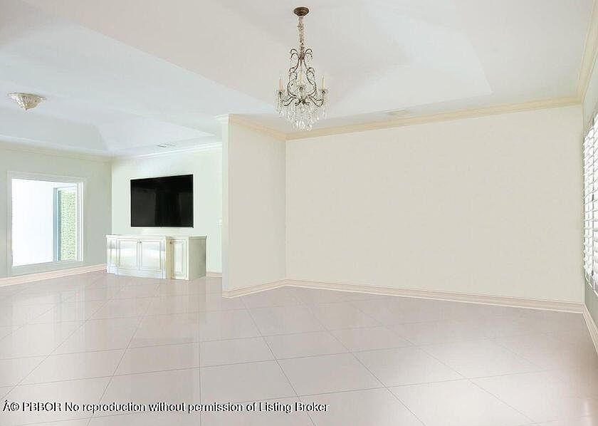 The image showcases a spacious living room with a neutral color palette, featuring light tile flooring and white walls. A chandelier hangs from the ceiling, adding a touch of elegance. A large television is mounted on the wall in an adjacent area, with a white cabinet underneath, suggesting an open-concept design.