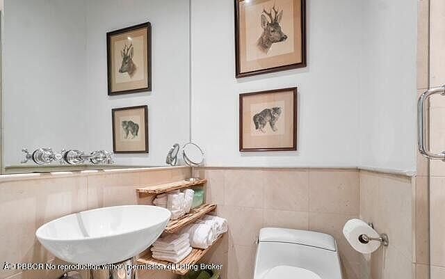 This is a well-lit guest bathroom featuring a modern vessel sink, a toilet, and decorative artwork. The walls are tiled in a neutral tone, and a wooden shelf provides storage for towels and toiletries. The overall impression is clean and stylish, creating a comfortable space for guests.
