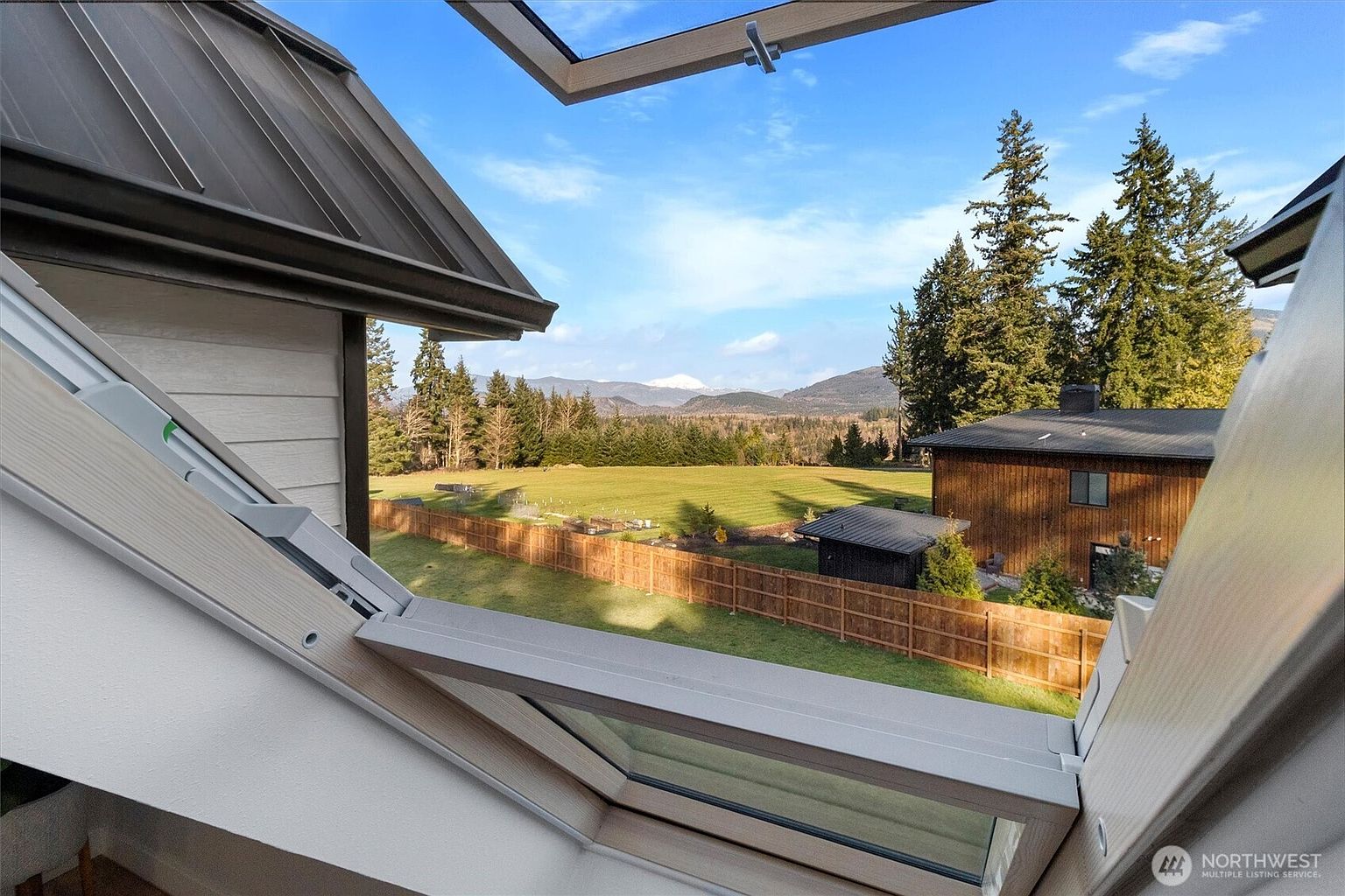 The image showcases a picturesque view of a well-maintained yard and garden, framed by an open skylight. A wooden fence encloses the lush green lawn, with trees and mountains visible in the background. A building is visible to the right of the frame, and the overall scene evokes a sense of tranquility and natural beauty.