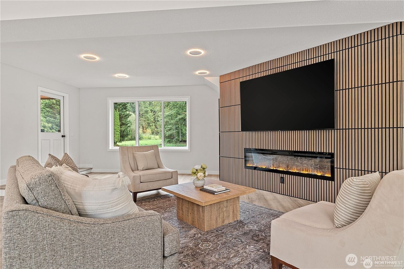 This is a bright and modern living room featuring a contemporary fireplace and TV wall. The room is furnished with comfortable seating, including a sofa and armchairs, arranged around a wooden coffee table. Large windows provide natural light and views of the outdoors, creating a welcoming and stylish space.