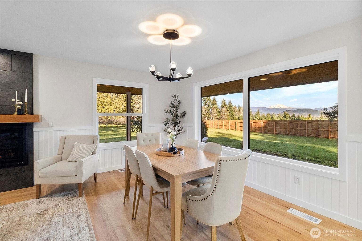 This is an interior shot of a dining room featuring a wooden table with six upholstered chairs. A modern chandelier hangs above the table, and large windows offer a view of the backyard and distant mountains. The room is decorated in neutral tones with white wainscoting, creating a bright and inviting space.
