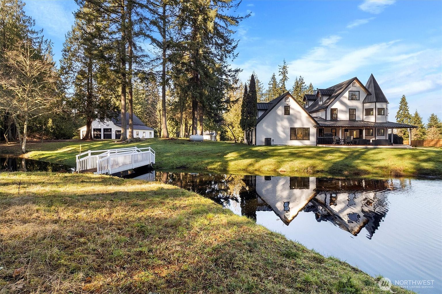 This image showcases the front exterior of a charming, modern farmhouse-style home with a picturesque pond reflecting its image. A white bridge leads to a smaller building on the left, while the main house features a combination of white siding, black-framed windows, and a striking turret. The well-manicured lawn and surrounding trees create a serene and inviting atmosphere.
