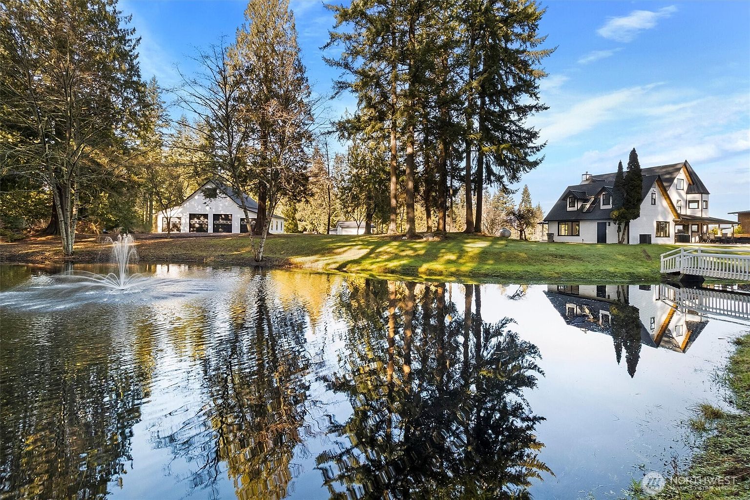 This picturesque exterior shot showcases a well-manicured yard featuring a serene pond with a fountain. The reflection of the surrounding trees and the house in the water adds depth and tranquility to the scene. The property includes a charming house and a separate garage, both contributing to the overall appeal of a peaceful, estate-like setting.