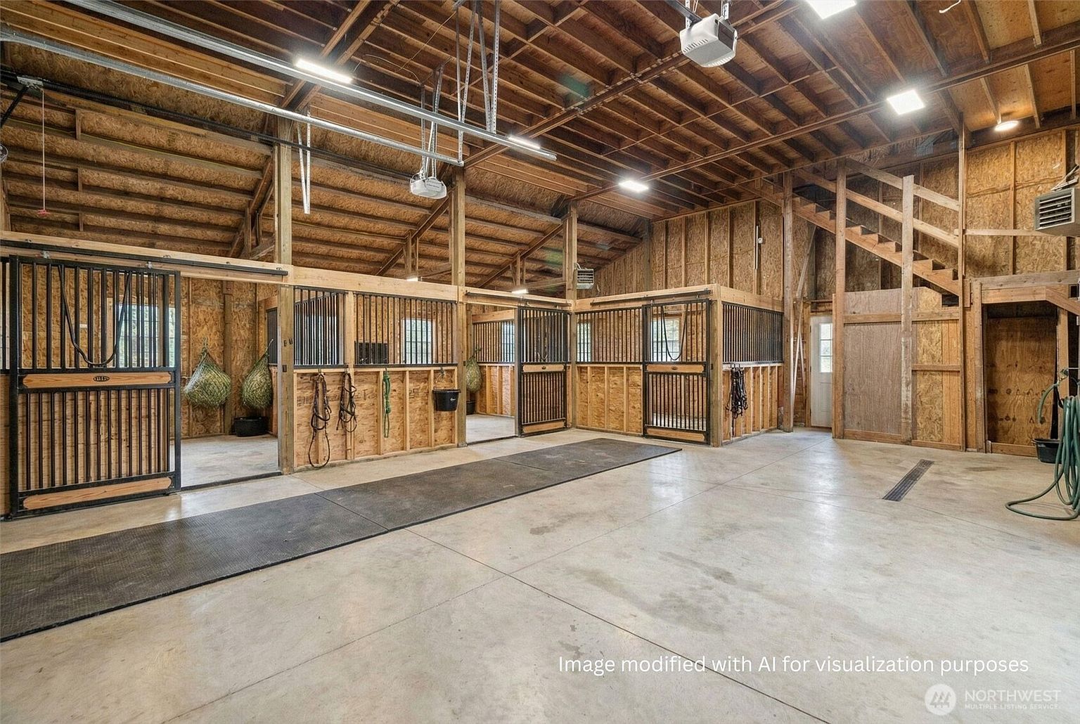 This image showcases the interior of a well-maintained barn, featuring wooden stalls with metal bars, a concrete floor, and a high, exposed wooden beam ceiling. The space is brightly lit with overhead lighting, and the stalls appear clean and functional, suggesting a well-cared-for equestrian facility. A staircase leads to an upper level, adding to the barn's functionality.