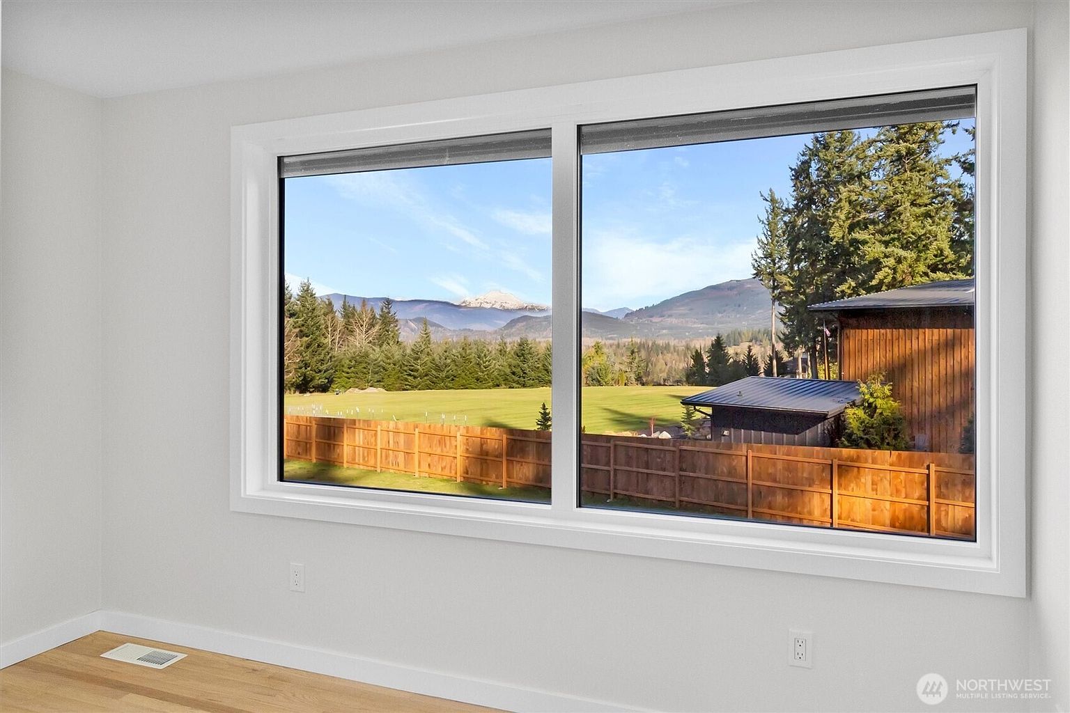 This interior shot showcases a bright room with a large window offering a scenic view of a green field, trees, and distant mountains under a clear sky. The room features light-colored walls and hardwood flooring, creating a clean and airy atmosphere, ideal for a bedroom or living space.