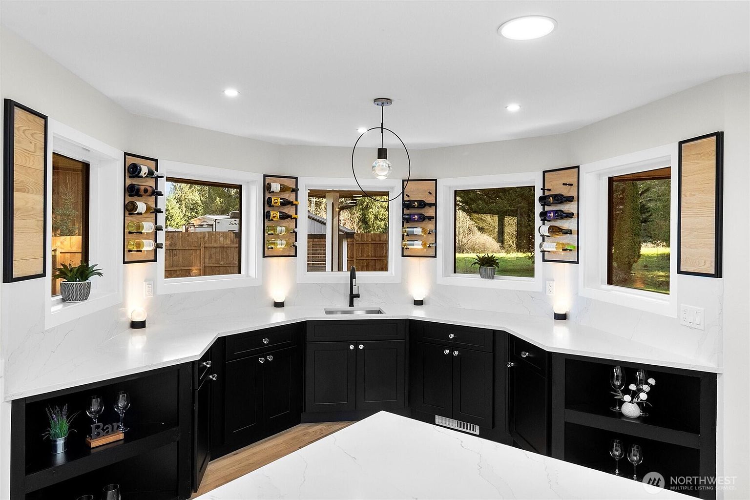 This is a well-lit kitchen with black cabinetry, white countertops, and a unique window design featuring built-in wine racks. The kitchen has a modern aesthetic with a focus on clean lines and functionality. The lighting fixtures and decor elements add a touch of elegance to the space.