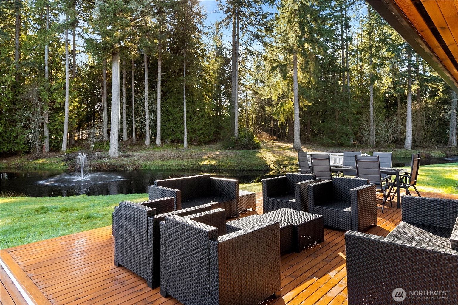 This image showcases a beautifully designed outdoor patio area, featuring modern wicker furniture arranged on a wooden deck. The patio overlooks a serene pond with a fountain, surrounded by lush greenery and tall trees, creating a peaceful and inviting atmosphere. A dining set is visible in the background, suggesting ample space for outdoor entertaining.