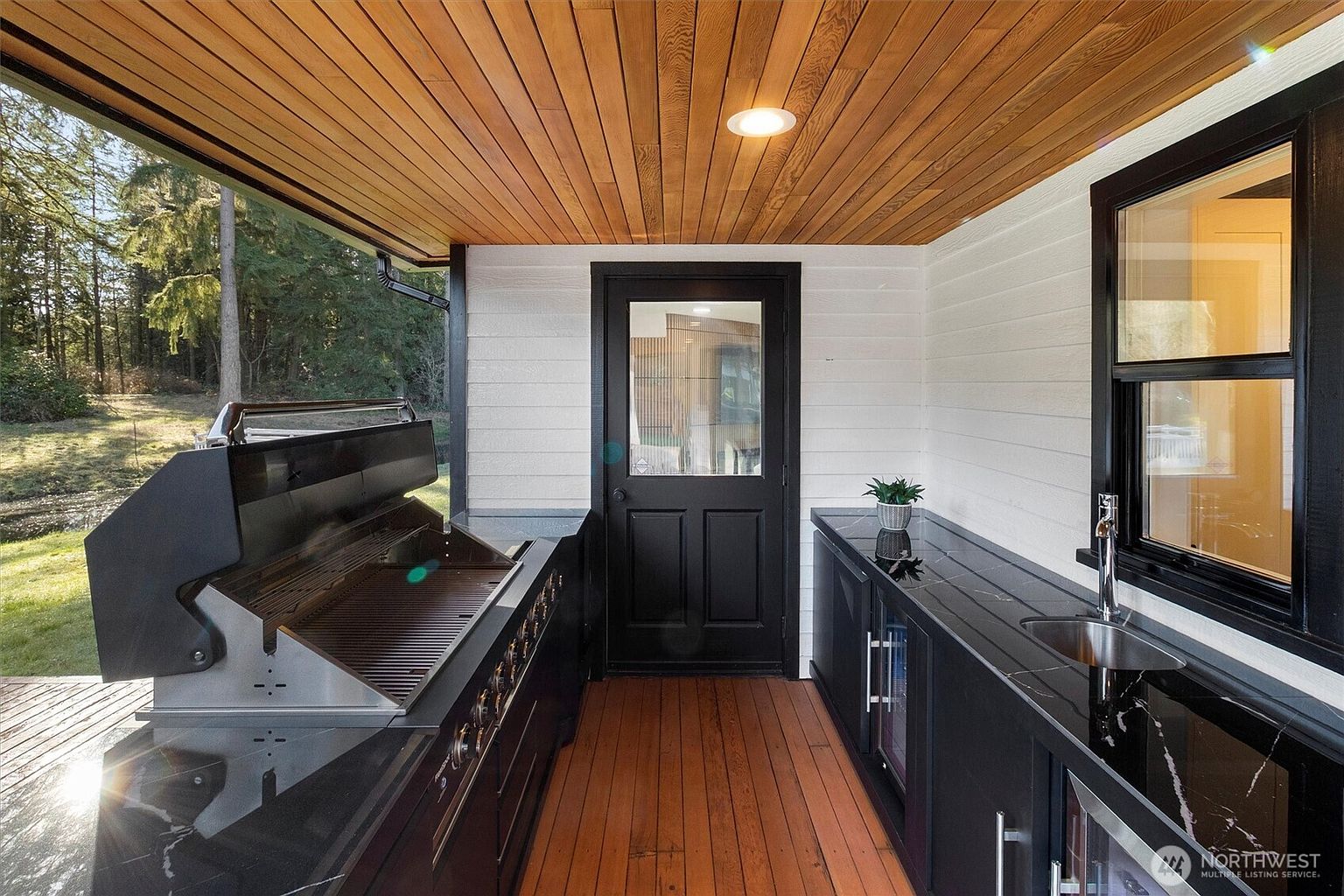 This image showcases an outdoor kitchen area on a patio or deck. The space features a built-in grill, dark cabinetry with a sleek countertop, a sink, and a beverage refrigerator. The ceiling is finished with wood planks, and the flooring is also wood, creating a warm and inviting atmosphere for outdoor entertaining.