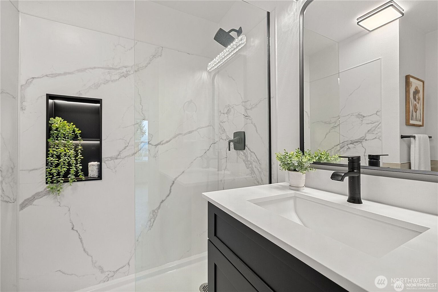 This is a well-lit primary bathroom featuring a modern design with white marble-patterned walls and a black vanity. The bathroom includes a glass-enclosed shower with a rainfall showerhead and a built-in niche with decorative plants. A sleek black faucet complements the white countertop and sink, while a large mirror reflects the light and space, enhancing the luxurious feel.