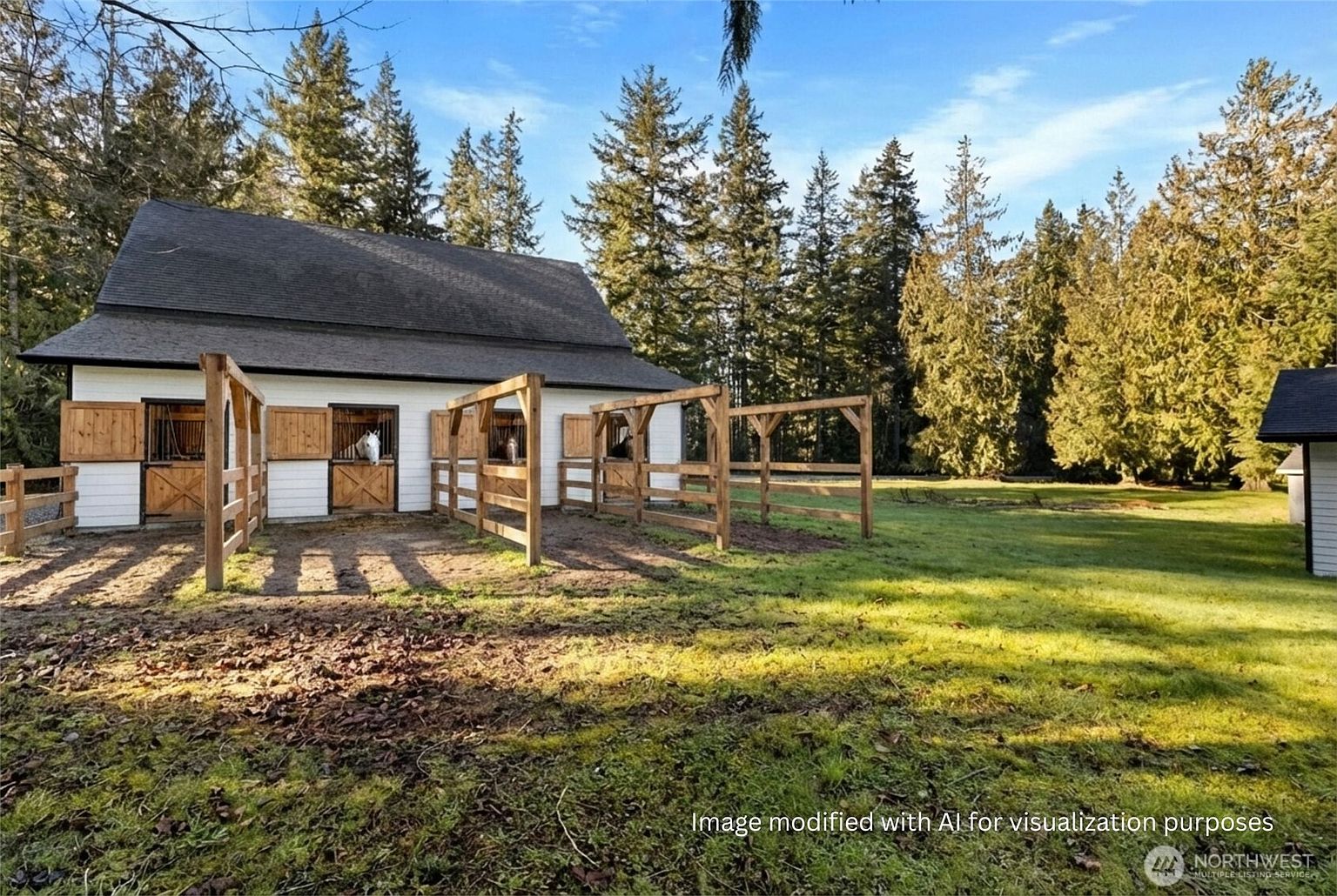 The image showcases a charming barn-style building with white siding and dark roofing, complemented by wooden fencing and a well-maintained grassy yard. The structure features stall openings with wooden doors, suggesting it could be used for horses or other livestock. The surrounding landscape includes mature trees, creating a serene and rural atmosphere.