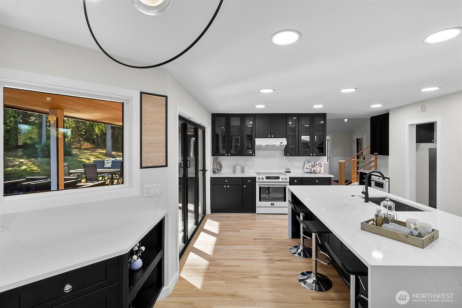 This is a bright and modern kitchen featuring black cabinetry with glass fronts, white countertops, and stainless steel appliances. A large island with bar seating anchors the space, while a window offers a view to the outside. The hardwood floors add warmth to the contemporary design.