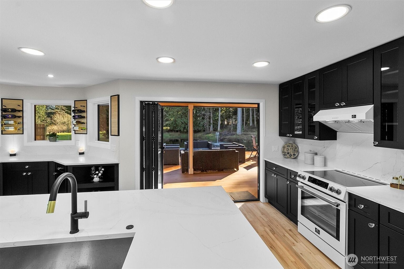 This is a bright, modern kitchen with black cabinetry and white countertops. A large island with a sink and a black faucet is in the foreground. The kitchen opens to an outdoor deck area through folding glass doors, offering a seamless transition to outdoor living.