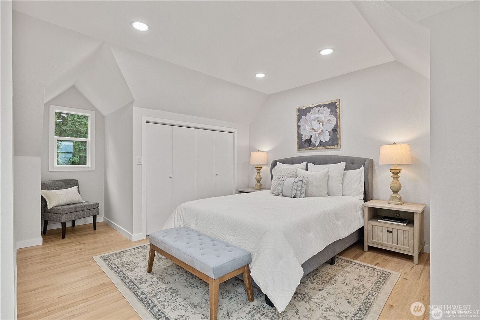 This is a bright and airy bedroom featuring a neutral color palette. The room includes a bed with white linens and decorative pillows, a bench at the foot of the bed, and matching nightstands with lamps. A window provides natural light, and a piece of artwork hangs above the bed, creating a serene and inviting atmosphere.