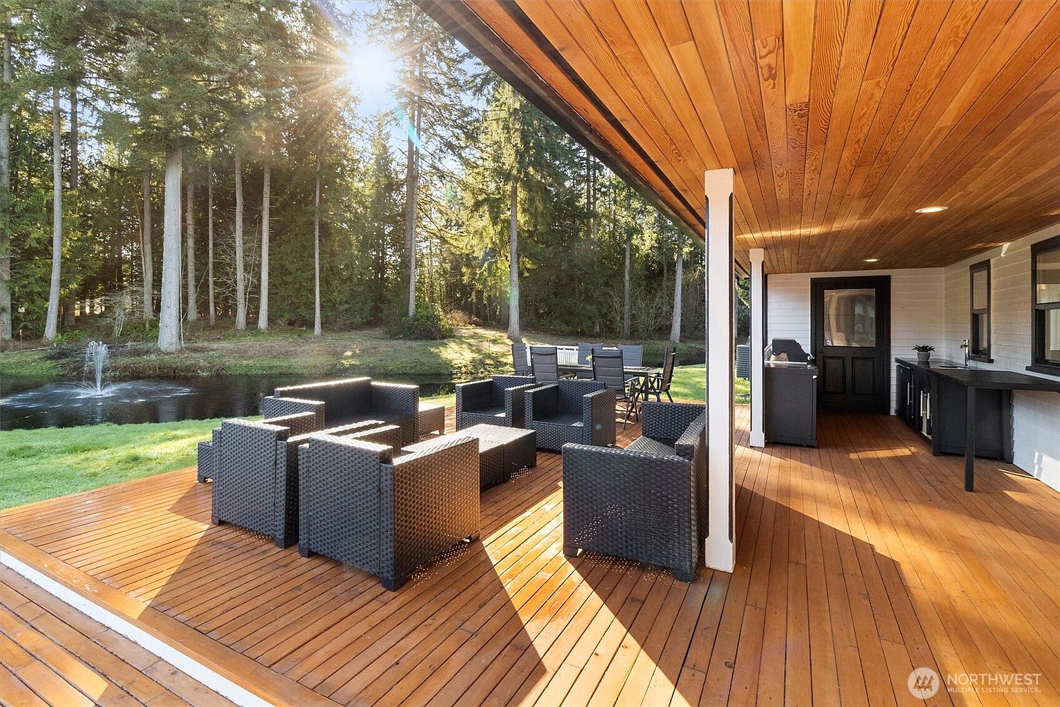 This image showcases a spacious outdoor patio area, featuring dark wicker furniture arranged for comfortable seating. The patio is constructed with warm-toned wooden planks, and a covered section provides shade, complete with a built-in outdoor kitchen area. Beyond the patio, a well-maintained lawn leads to a serene pond with a fountain, all surrounded by lush trees, creating a tranquil and inviting outdoor living space.