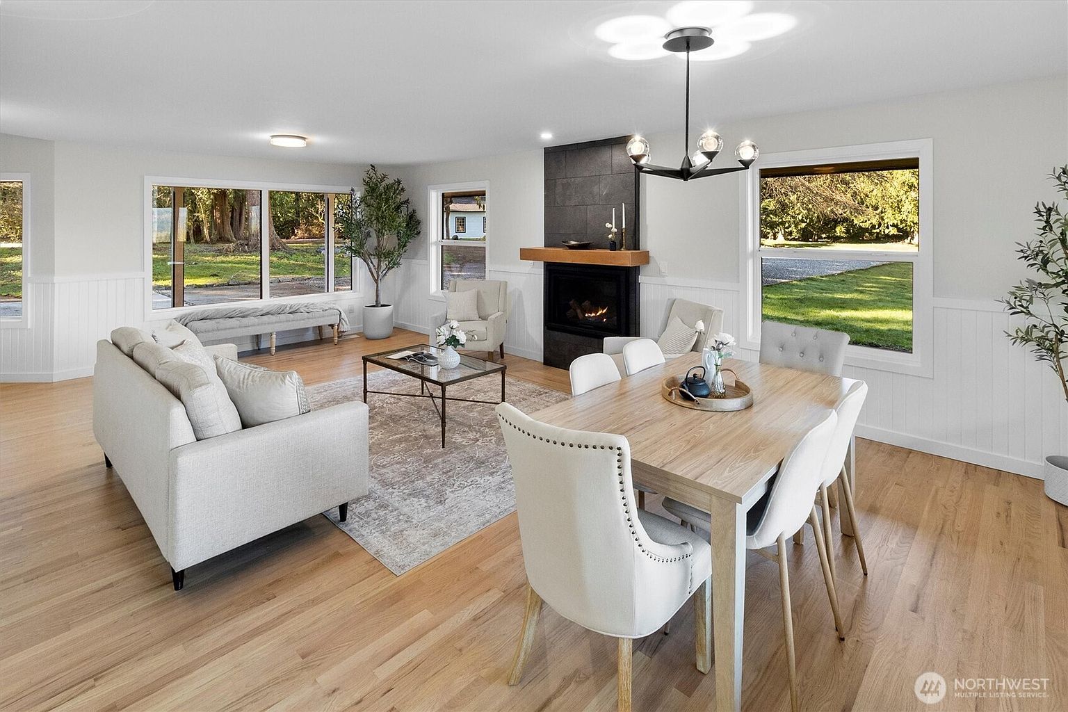 This is a bright and airy living and dining area featuring hardwood floors, white wainscoting, and large windows that offer ample natural light. The living room includes a comfortable sofa, a glass coffee table, and a decorative tree, while the dining area showcases a wooden table with upholstered chairs and a modern chandelier. A fireplace with a dark tile surround adds a focal point to the space.