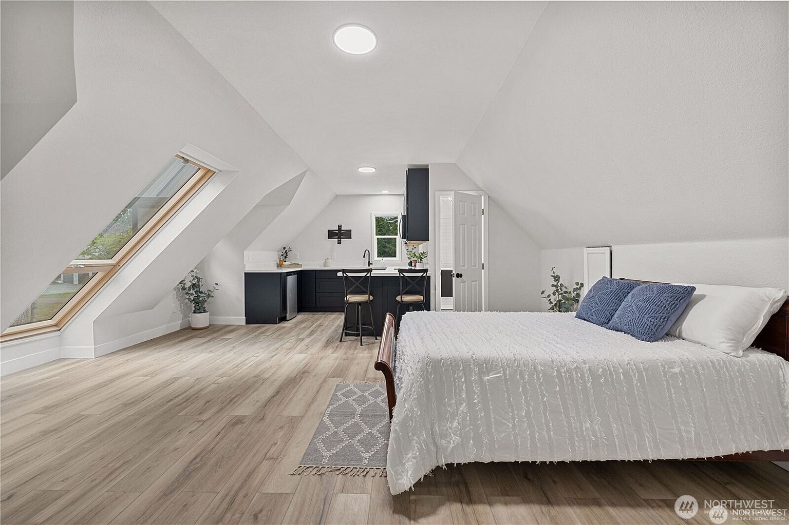 This is an interior shot of a bedroom featuring a kitchenette. The room has a bed with white linens and blue pillows, light wood flooring, and a small kitchen area with dark cabinets and bar stools. A skylight and a window provide natural light, enhancing the bright and airy feel of the space.