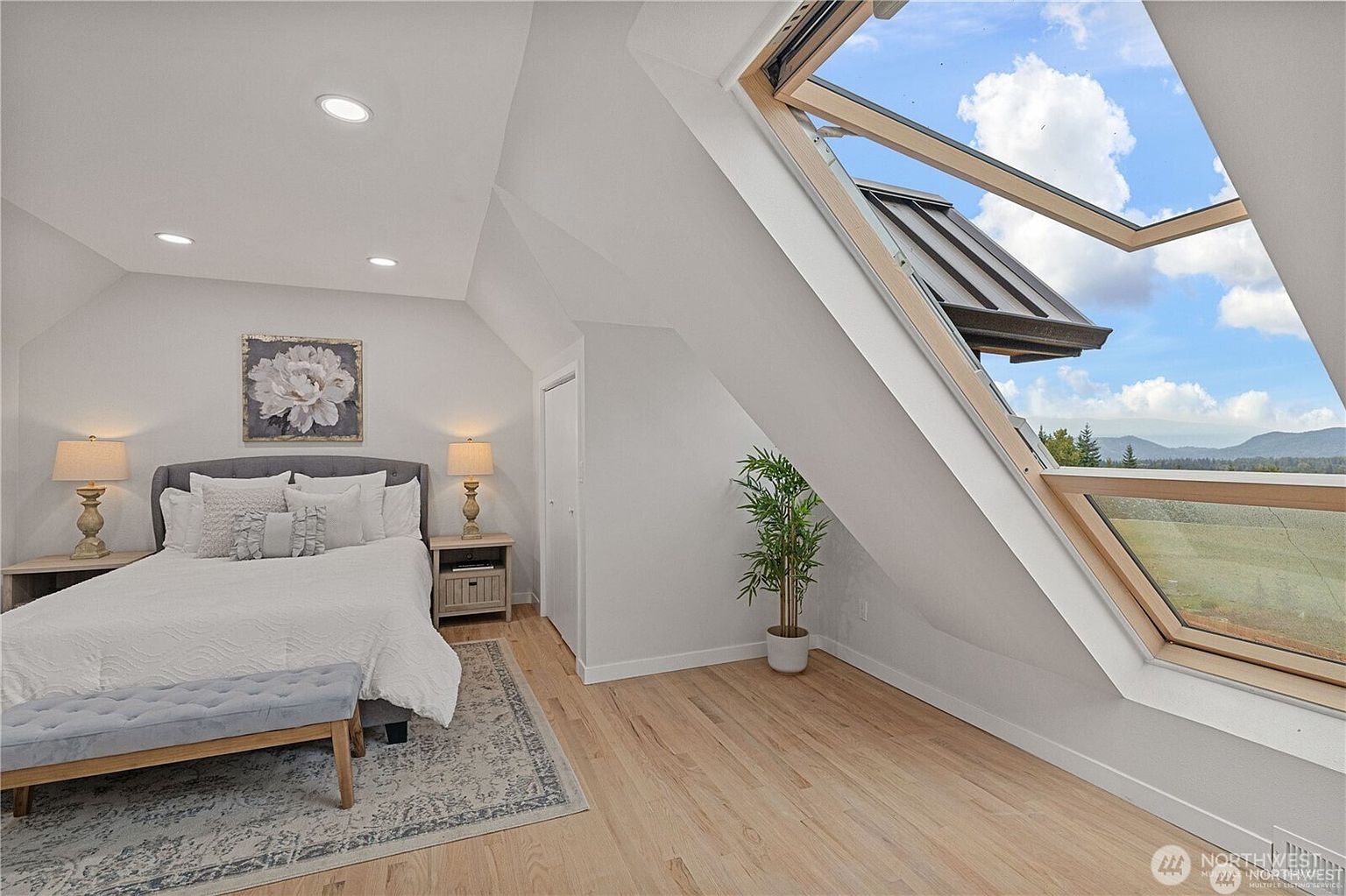 This is a bright and airy bedroom featuring a large skylight with a view of the sky and distant mountains. The room is furnished with a bed, side tables with lamps, a bench, and a decorative plant, creating a cozy and inviting atmosphere. The hardwood floors and neutral color palette enhance the sense of spaciousness and tranquility.