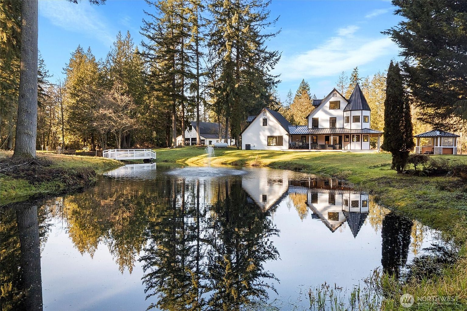 This image showcases the front exterior of a charming white farmhouse-style home with black accents, reflected beautifully in a serene pond. A white bridge leads to the property, and mature trees surround the house, creating a picturesque and inviting setting. The overall impression is one of tranquility and classic elegance.