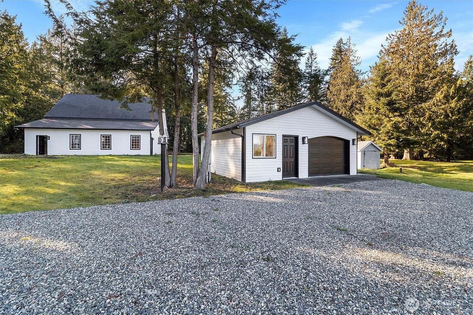 This is a front exterior view of a property featuring a white house with a dark roof and a detached garage. The garage, also white with dark trim, has a brown garage door and a side entrance. A gravel driveway leads up to the garage, and the property is surrounded by trees and a well-maintained lawn, creating a serene and private setting.